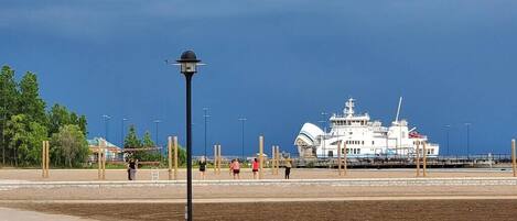 A waterfront area with a large white cruise ship docked, a promenade with people walking, and a street lamp in the foreground.