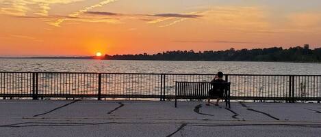 A sunset view from a waterfront promenade with a bench and a fence.