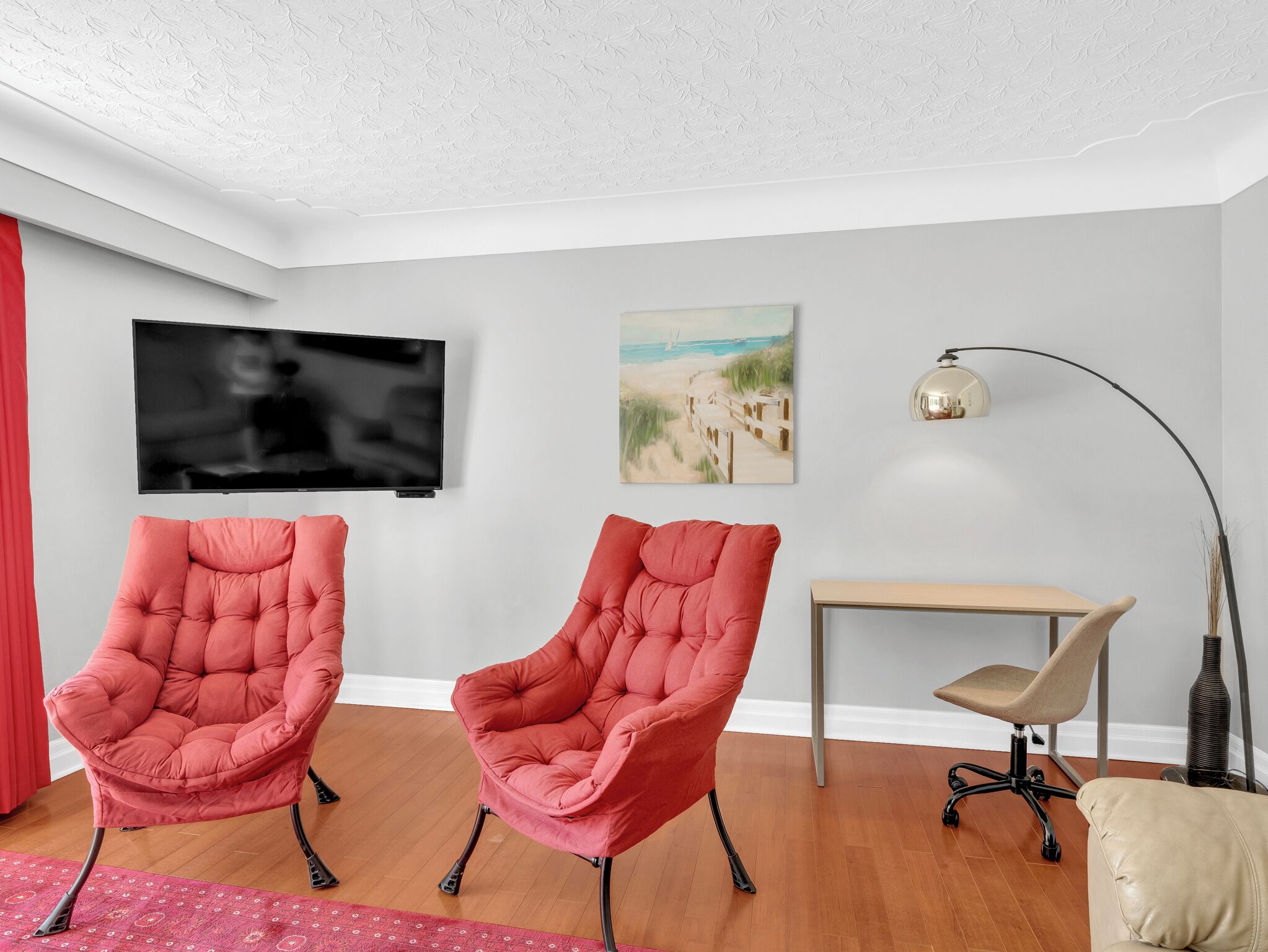 A modern living room with two red armchairs, a TV, a painting, a desk, and a lamp.
