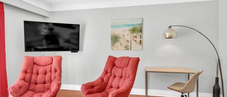 A modern living room with two red armchairs, a TV, a painting, a desk, and a lamp.