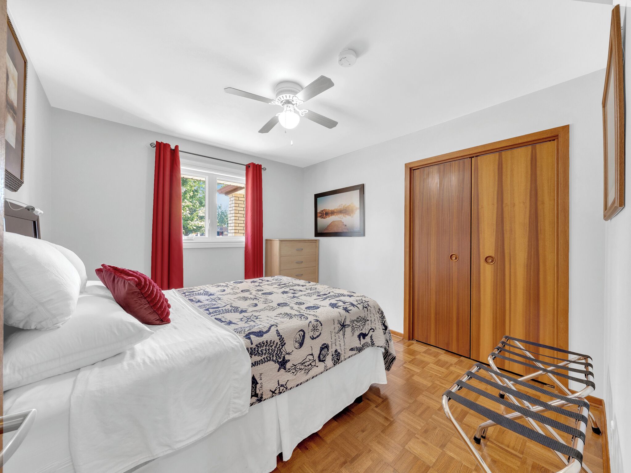 A bedroom with a bed, a wooden wardrobe, a ceiling fan, and a window with red curtains.