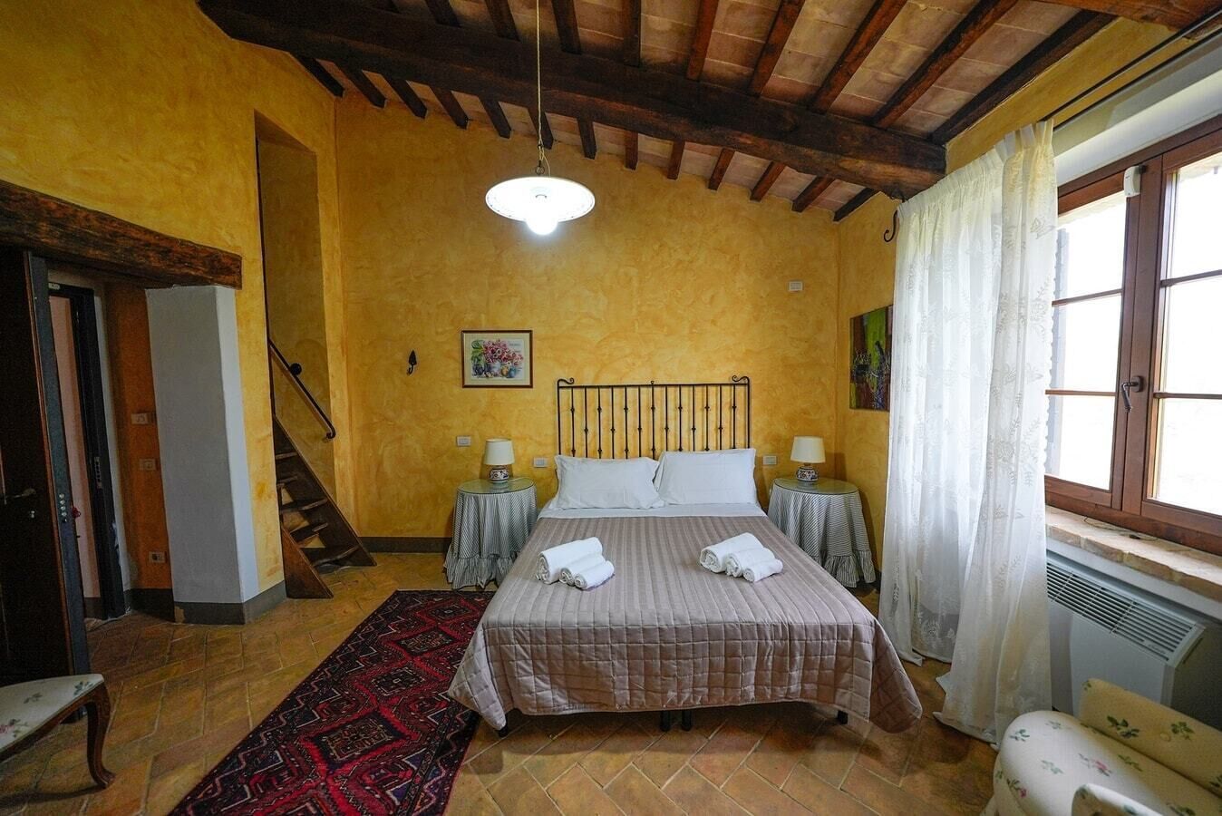 A bedroom with a bed, two bedside tables, a window with curtains, a radiator, and a staircase.