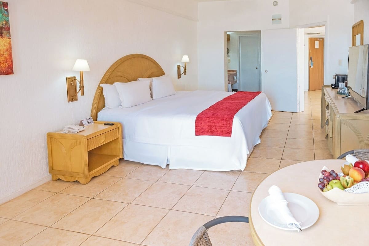 Habitación de hotel con una cama grande, mesitas de noche, un escritorio y un área de comedor con fruta.