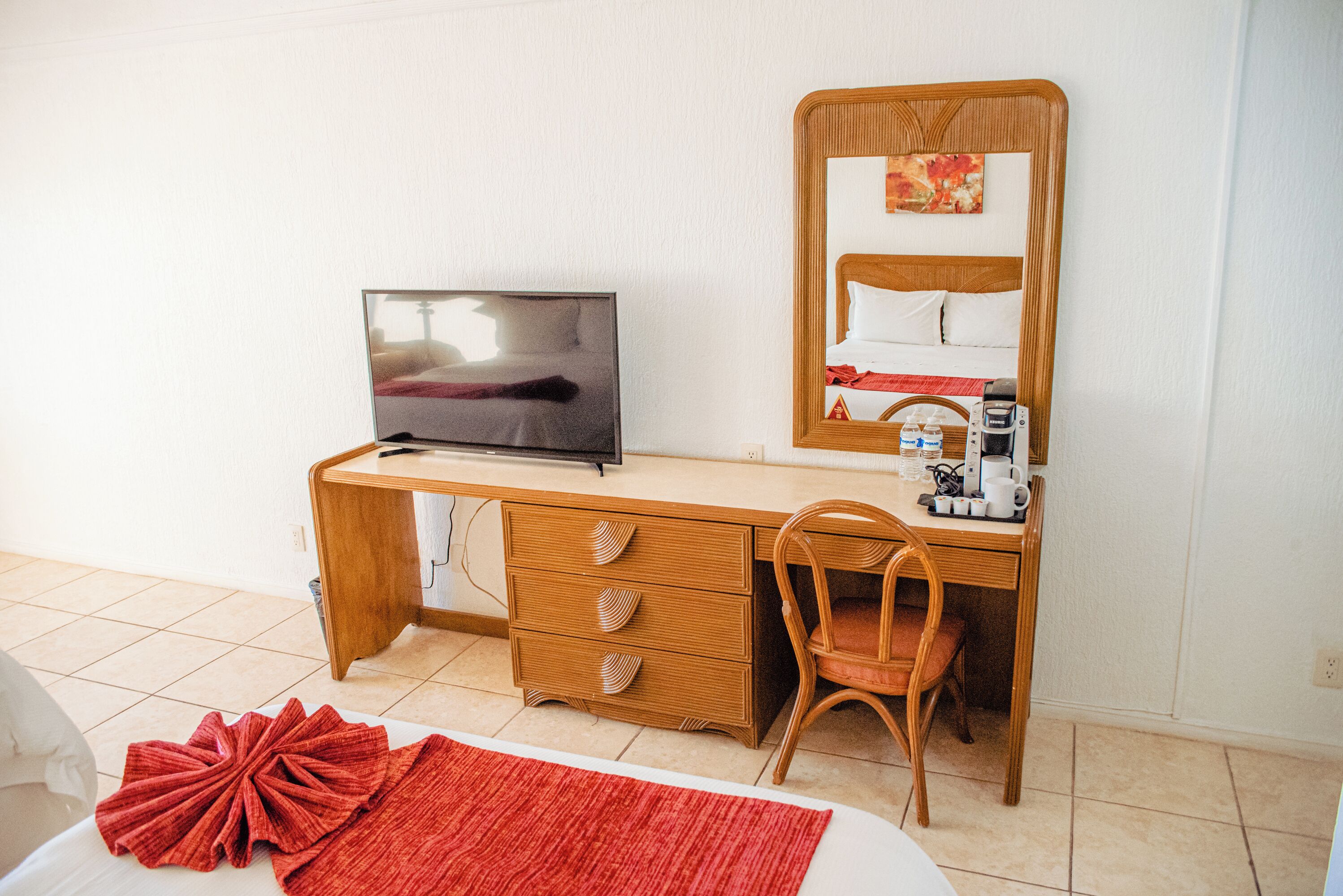 A hotel room with a wooden desk, a television, a bed with a red rug, a chair, and a mirror.