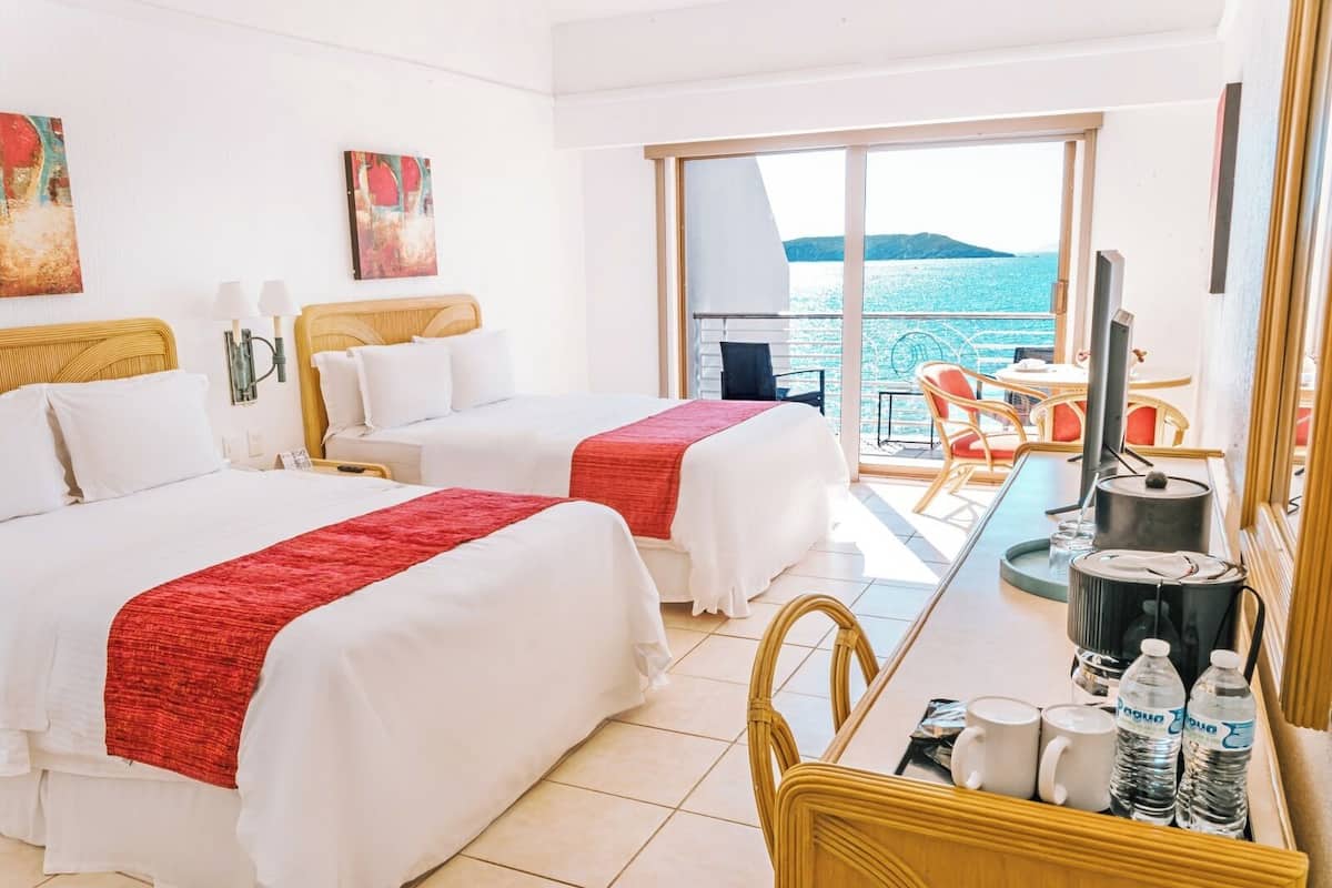 Una habitación de hotel con dos camas, un balcón con vistas al mar y un comedor con mesa y sillas.