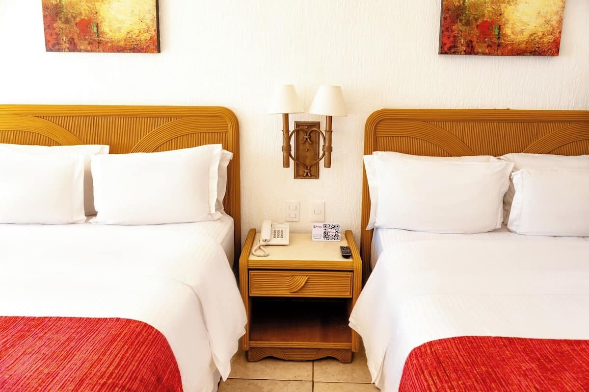 Habitación de hotel con dos camas, cada una con ropa de cama blanca y mantas rojas. Entre las camas hay una mesita de noche de madera con una lámpara, un teléfono y un cartel con un código QR.