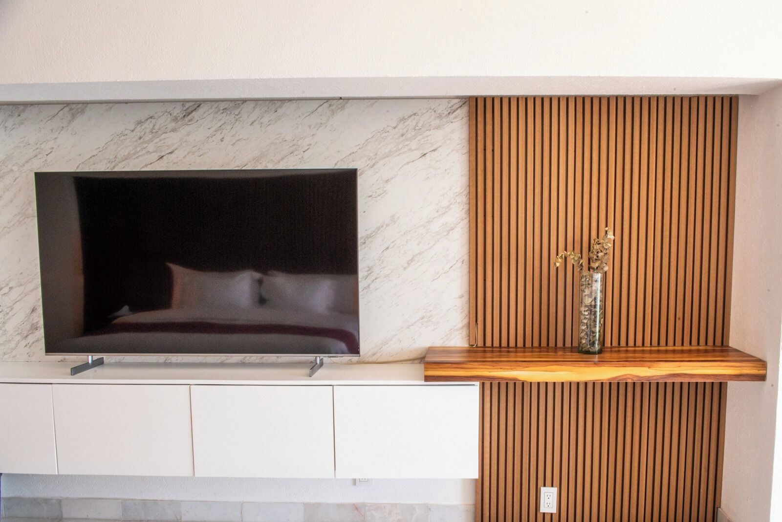 A modern living room with a large flat-screen TV mounted on a marble wall, a wooden slatted panel, and a small shelf with a vase.