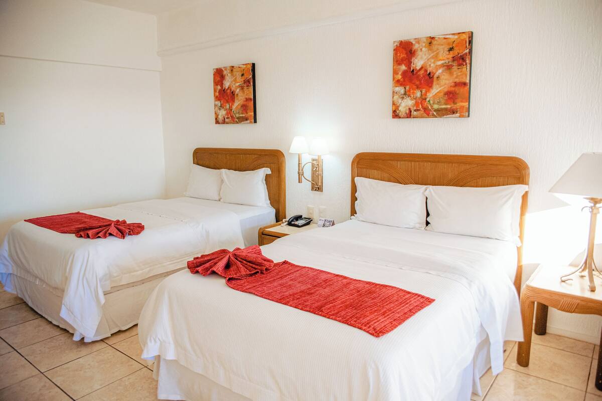 Habitación de hotel con dos camas, cada una con ropa blanca y detalles rojos, una mesita de noche con lámpara y arte abstracto en las paredes.
