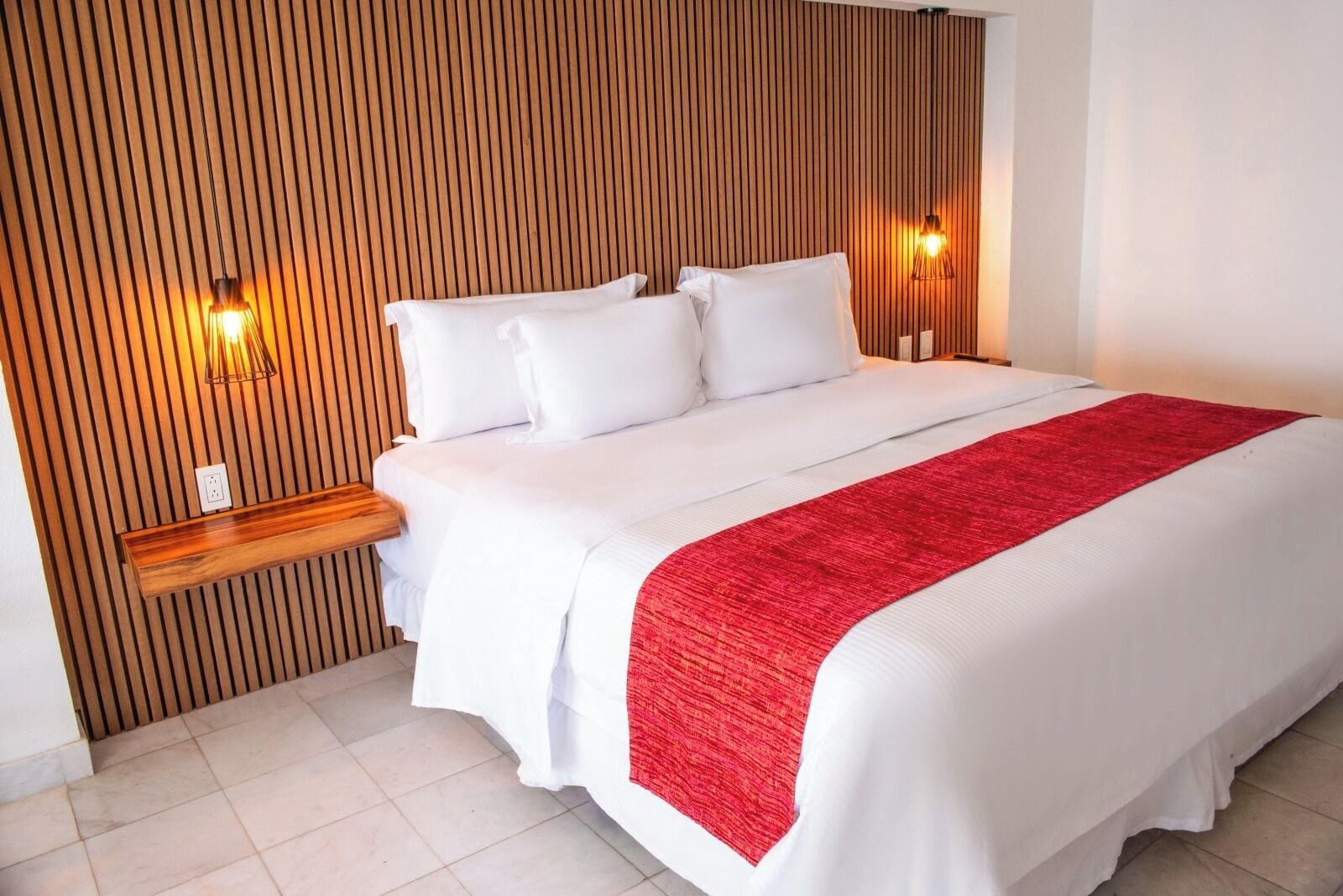 Een net opgemaakt bed met witte lakens en een rode plaids, een houten lattenbalken hoofdbord en twee wandlampen voor het lezen.