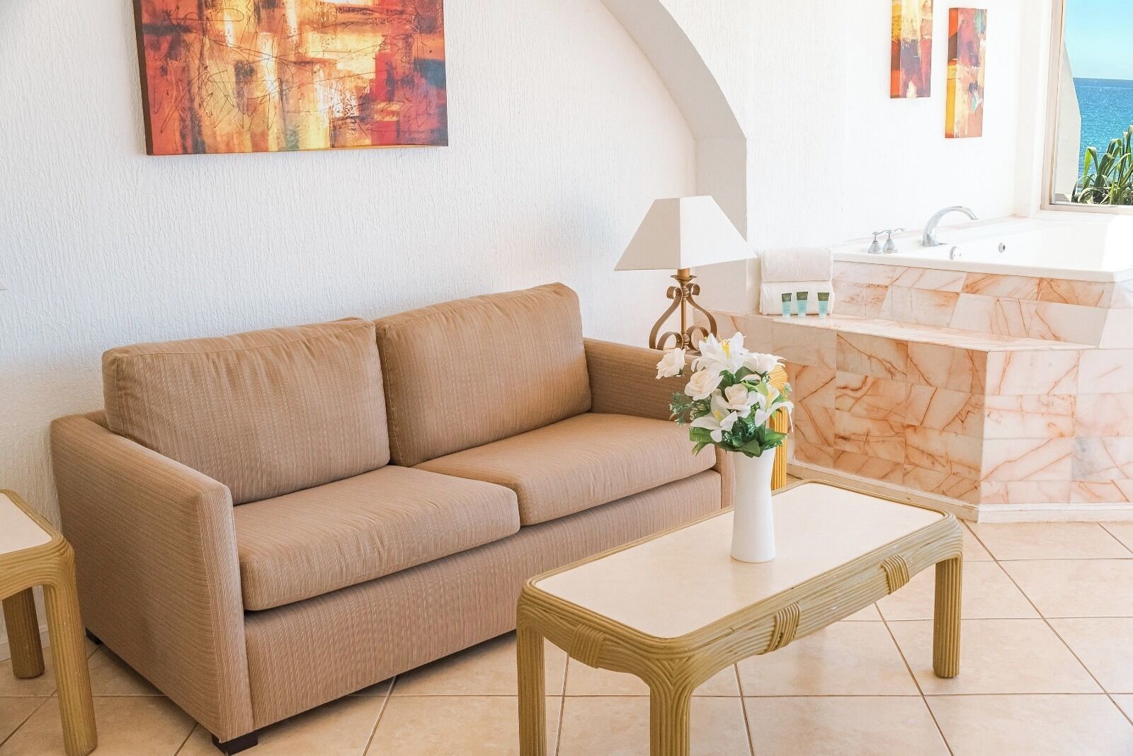 Een beige bank, een witte vaas met bloemen, en een badkamer met een marmeren wastafel en bad.