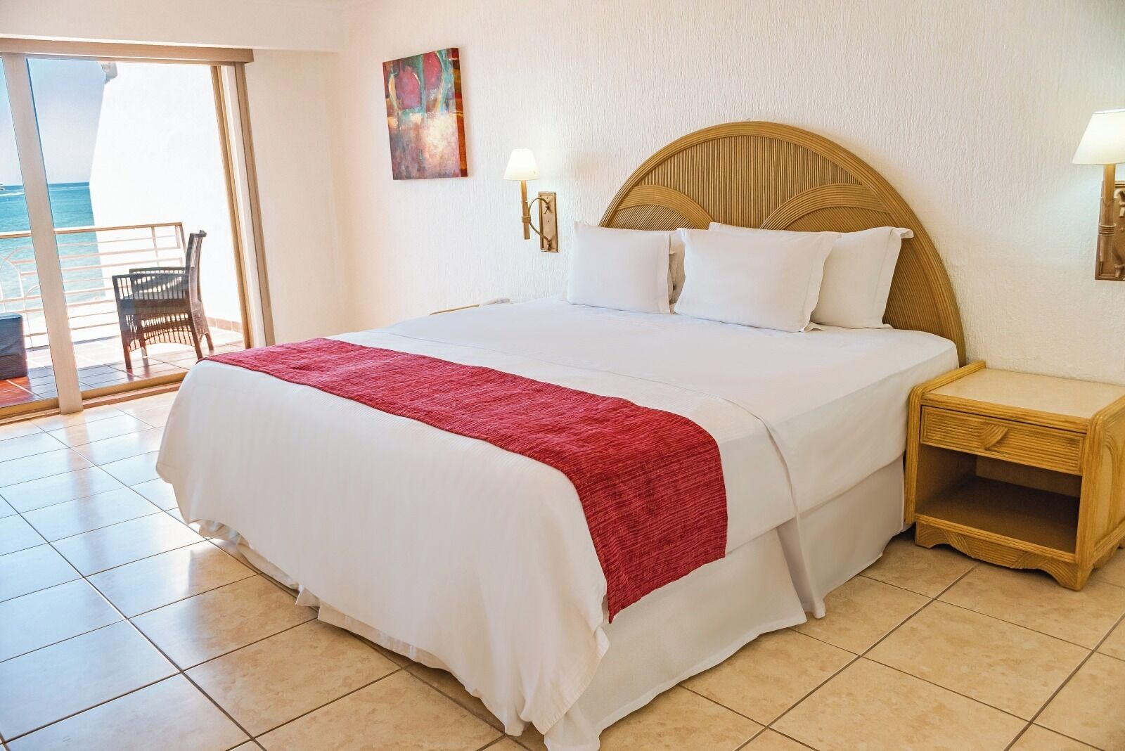 Habitación de hotel con una cama grande, una mesita de noche de madera, una manta roja, un cuadro en la pared y vista al océano a través de una puerta corredera.