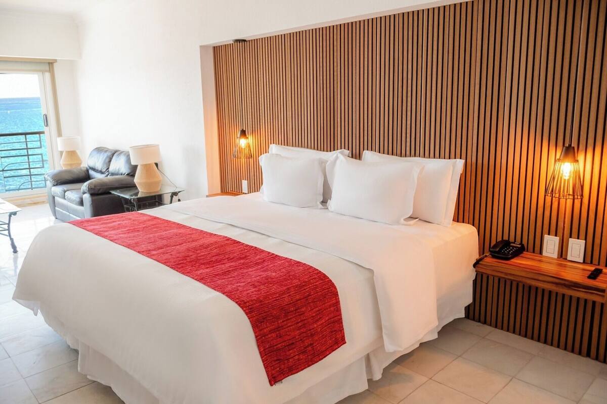 Habitación de hotel con una cama grande, cabecera de madera, manta roja y sofá de cuero, con vista al mar.