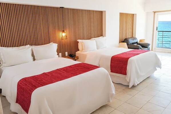 Habitación de hotel con dos camas, cabecera de madera con listones, mesita de noche, lámpara y vista al mar.