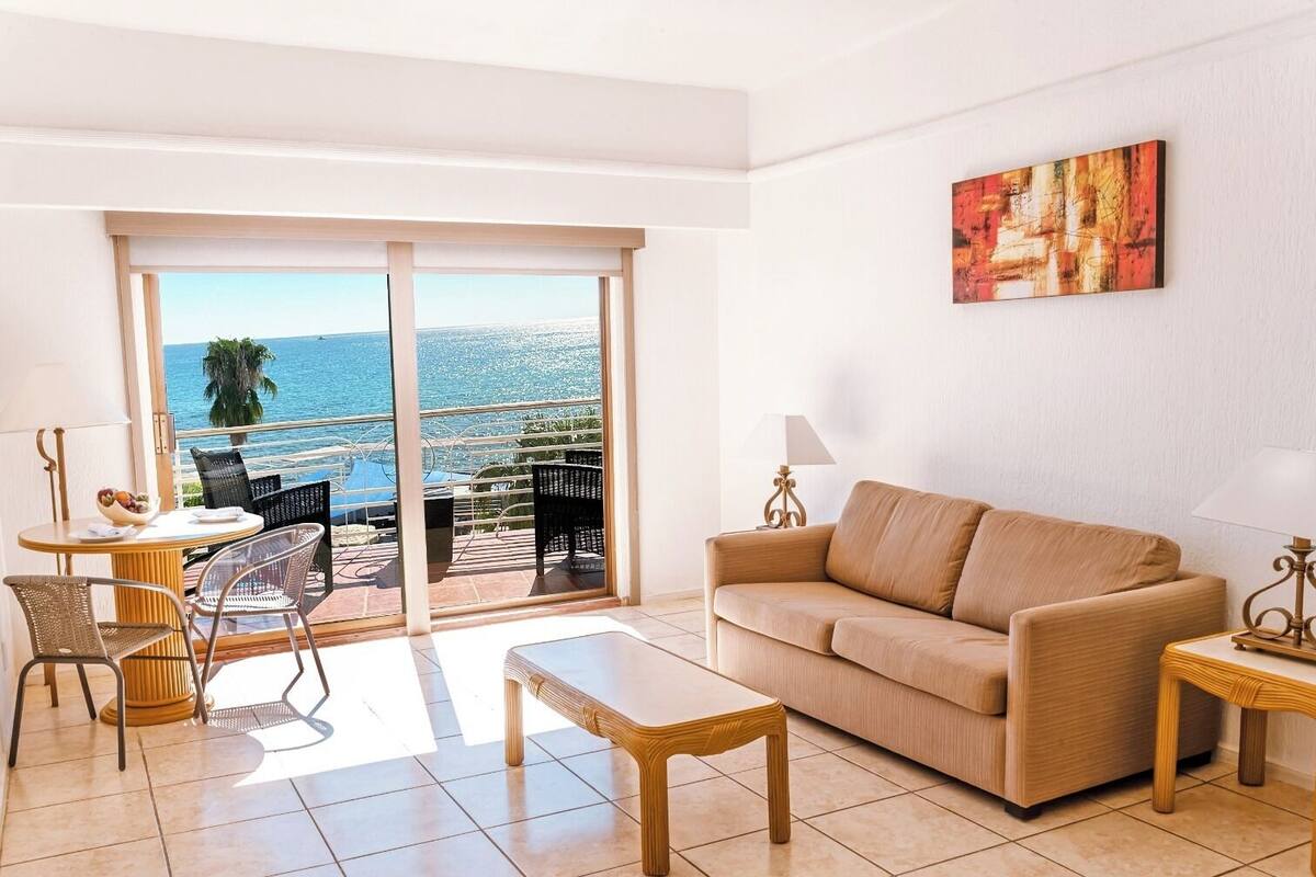 Una sala de estar con un sofá, una mesa de centro y un comedor con sillas y una mesa. Hay un ventanal con vista al mar.