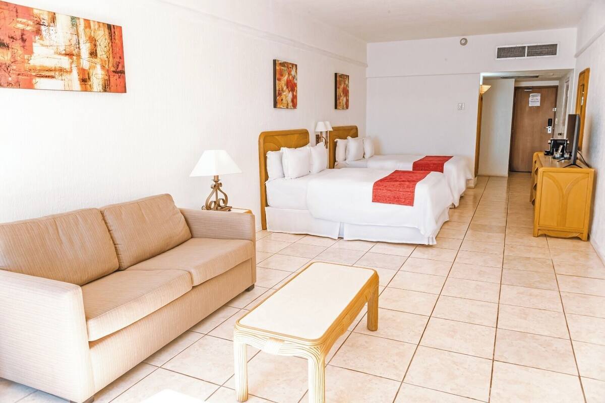 Una habitación de hotel con un sofá color beige, una mesita, una cama doble con ropa blanca y una manta roja, y dos cuadros en la pared.