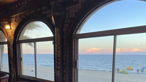 A beachside restaurant with large windows offering a view of the sea and beach.