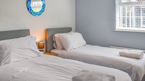 Hotellihuone, jossa on kaksi sänkyä, yöpöytä lampulla, sininen mosaiikkikuvioinen seinäkoriste ja ikkuna, jossa on sälekaihtimet.