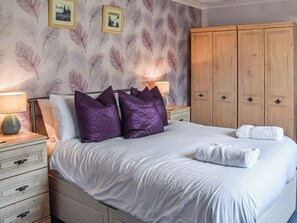 Ein ordentlich bezogenes Bett mit weißen Leinen und violetten Kissen, ein Holzschrank und eine Wand mit Faser-Muster-Tapete.