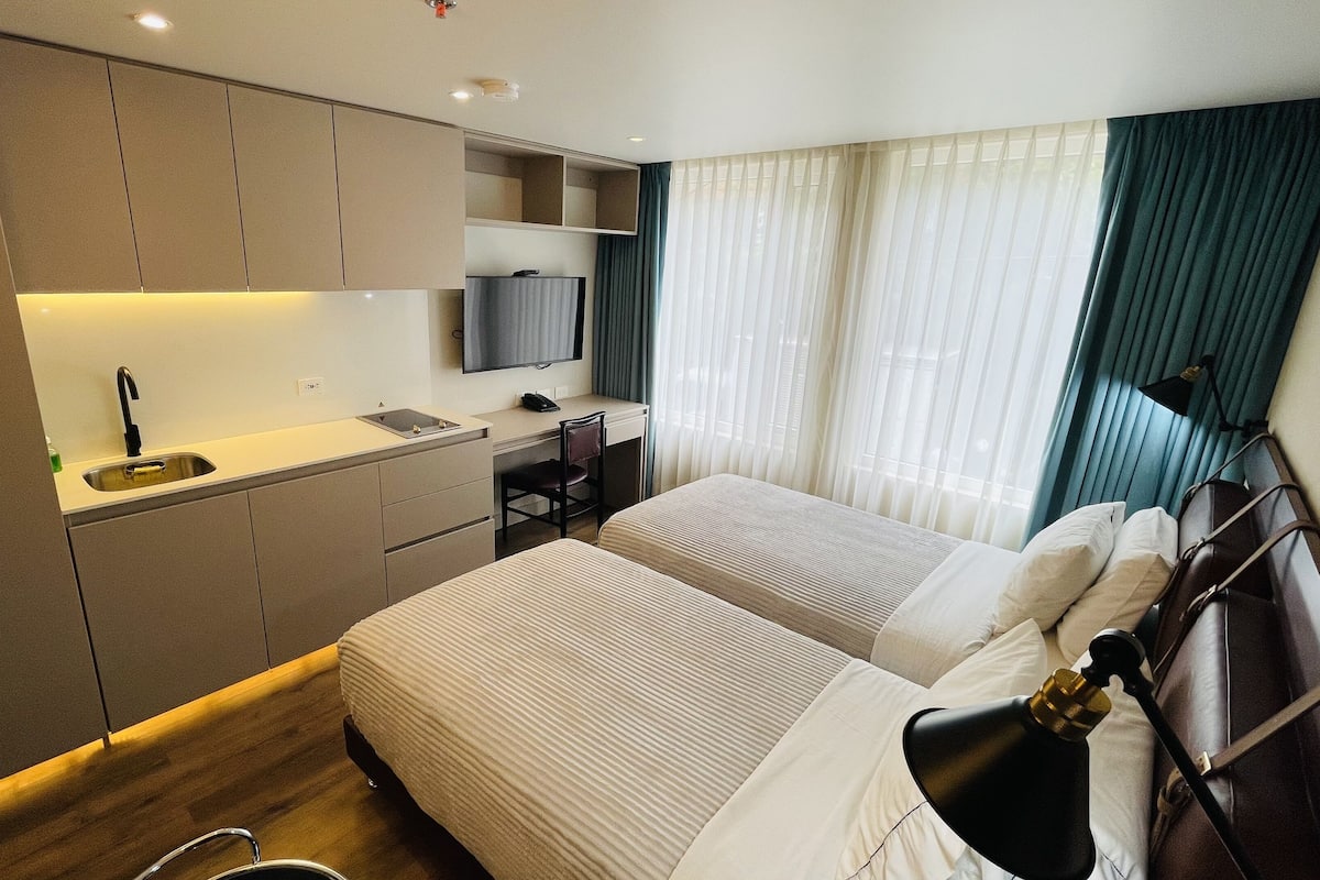 Habitación de hotel con dos camas, un escritorio con una silla, un televisor y una ventana con cortinas.