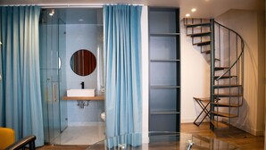 Un baño moderno con cabina de ducha de vidrio, un espejo redondo, un lavabo blanco y una escalera de caracol.