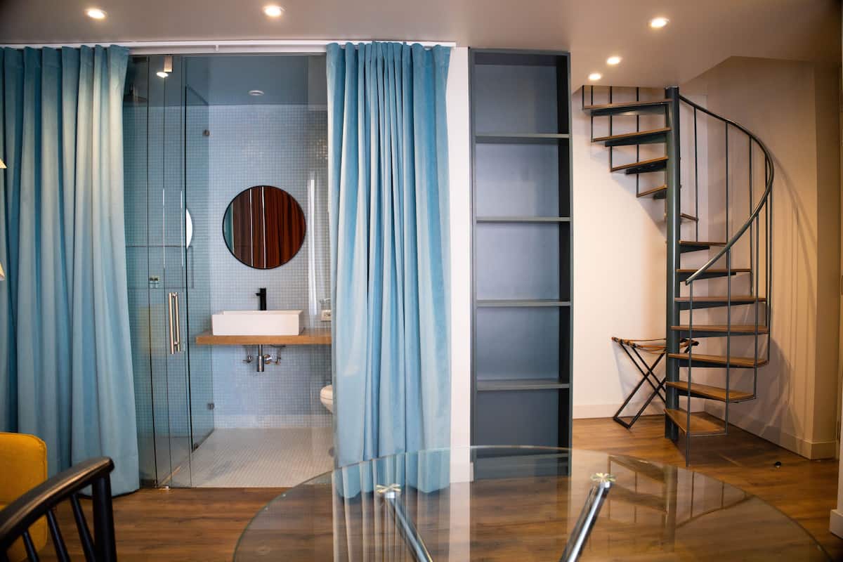 Un baño moderno con cabina de ducha de vidrio, un espejo redondo, un lavabo blanco y una escalera de caracol.