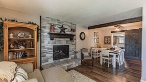 Una sala de estar con chimenea de piedra, una estantería, un comedor con mesa y sillas, y un pasillo que conduce a otra habitación.