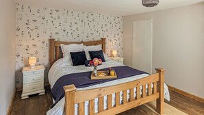 A bedroom with a wooden bed, white nightstands, a floral wallpaper, and a dark blue rug. - A nature lovers delight. (Ludham)