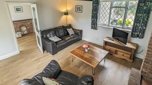 A living room with a black leather sofa, a wooden coffee table with a vase of flowers, a television on a wooden stand, and a fireplace with a 'Fresh Eggs' sign. - A nature lovers delight. (Ludham)