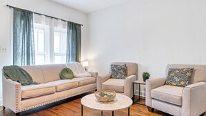 Een woonkamer met een beige bank, twee fauteuils, een ronde salontafel en een raam met gordijnen.