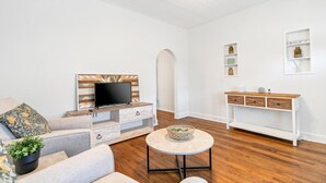 Een woonkamer met een bank, een salontafel, een televisie op een houten standaard en een witte console met planken.