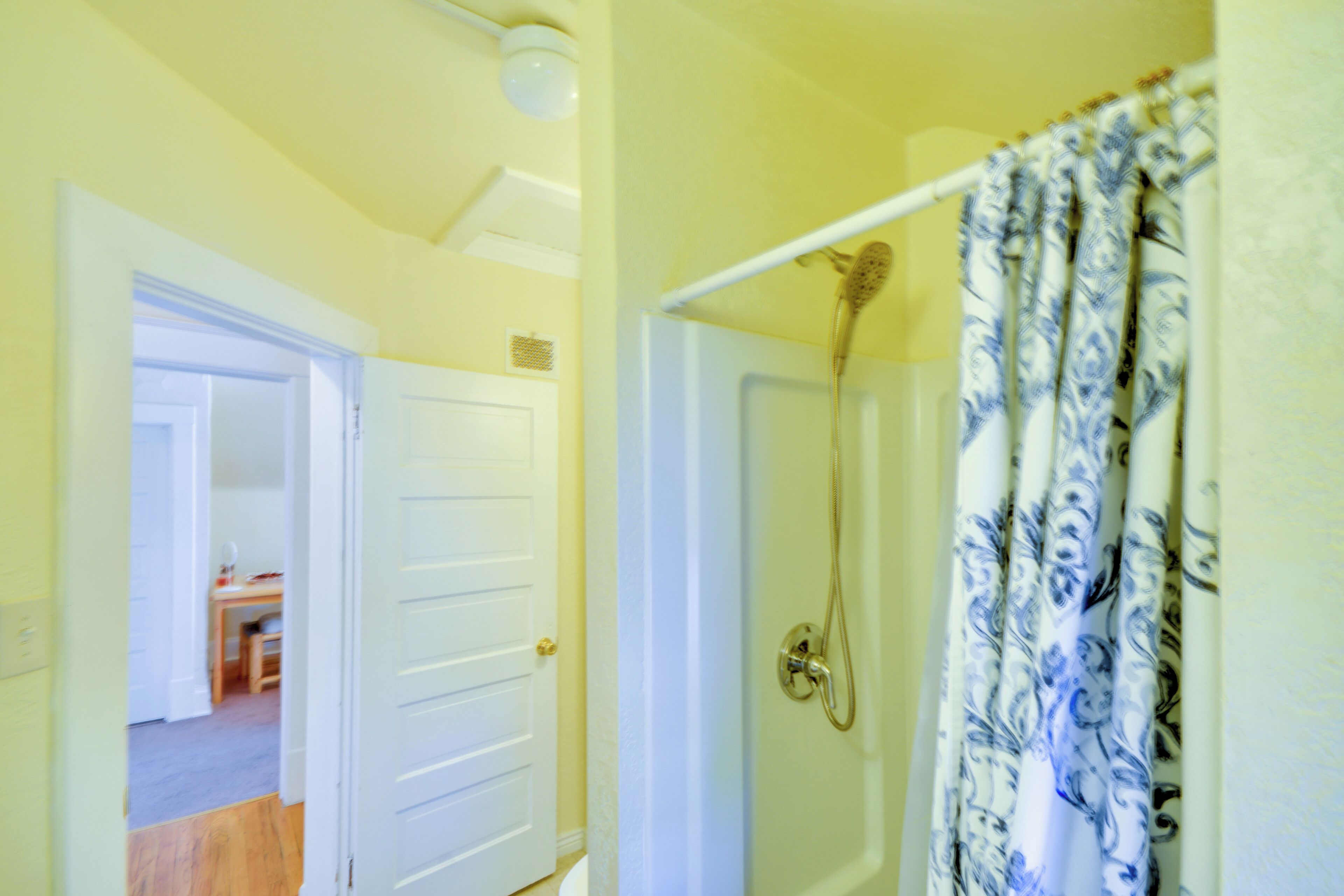 A bathroom with a shower, a door leading to a room, and a curtain with a floral pattern.