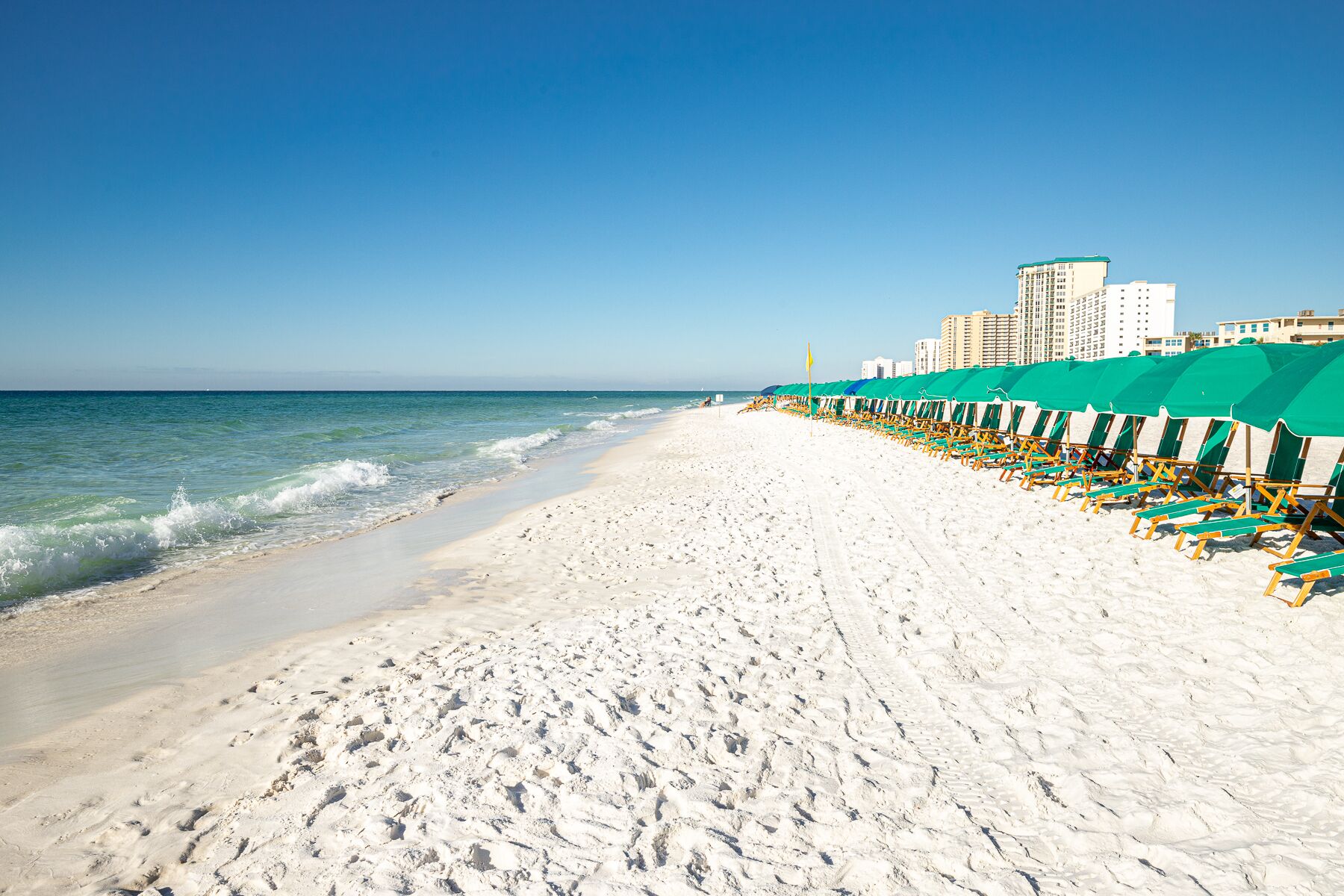 Ein Strand mit Reihen aus grünen Sonnenschirmen und Liegestühlen, ein sandiger Uferstreifen und ein klarer, blauer Himmel.