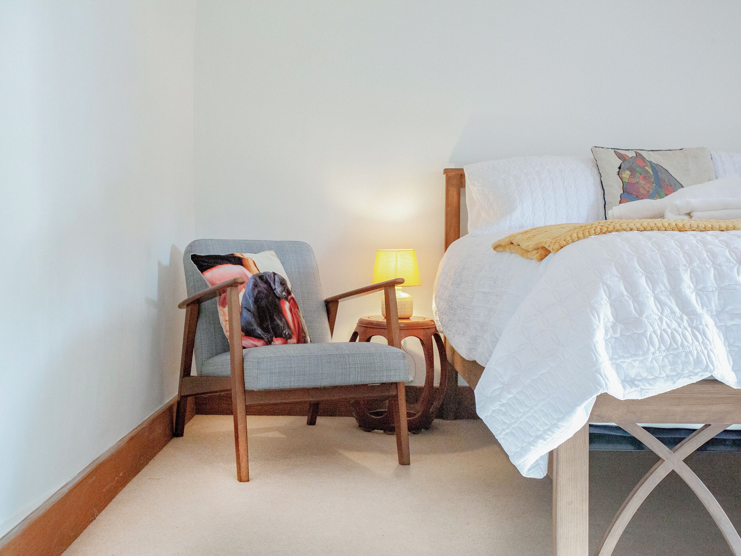 A bedroom with a bed, a chair with a pillow, a nightstand with a lamp, and a wall with a painting.