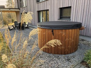 A wooden hot tub with a black cover, surrounded by gravel and plants, in front of a modern building with vertical siding.