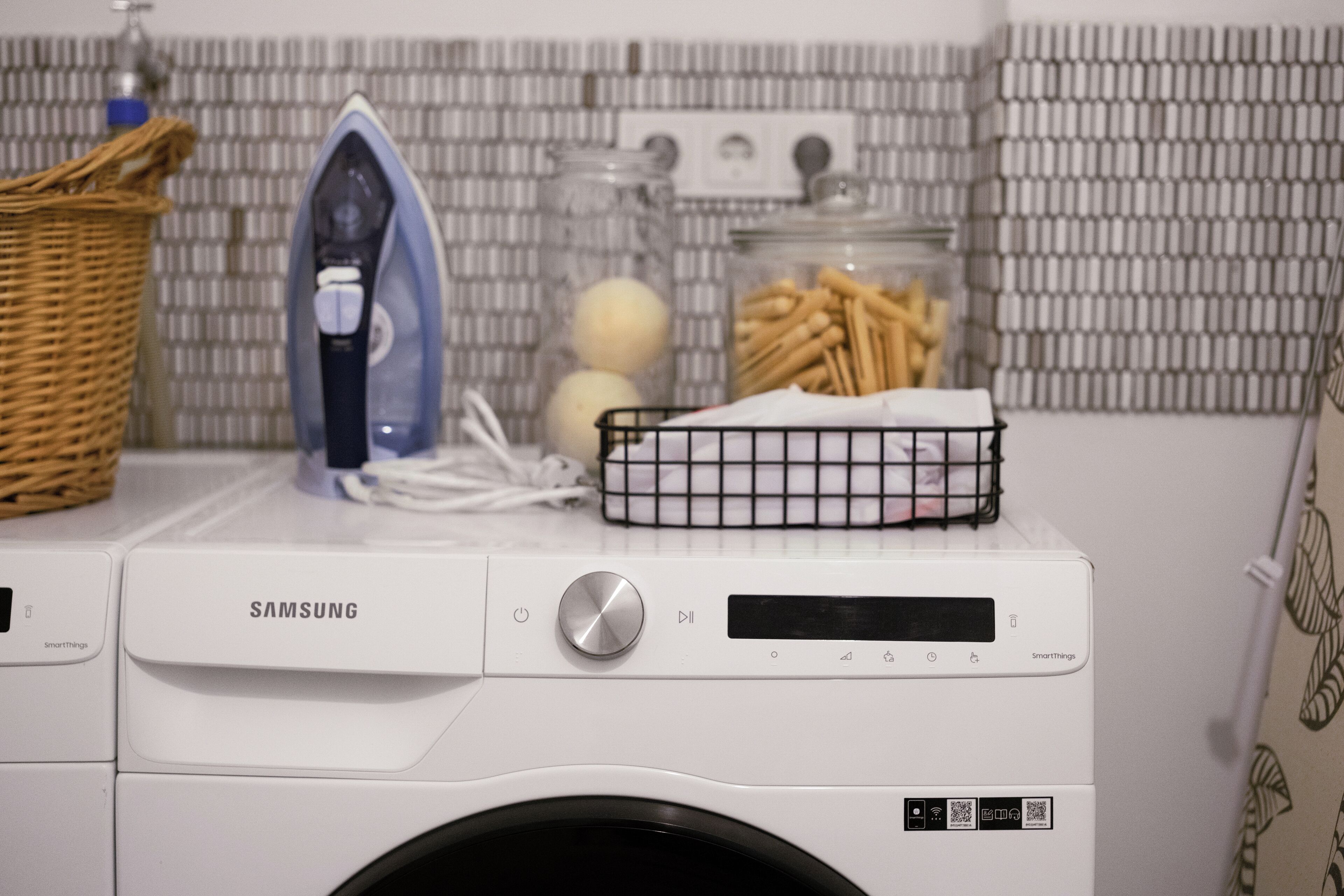 En Samsung SmartThings-tvättmaskin i ett tvättrum med en korg, ett strykjärn och en burk med diverse föremål.