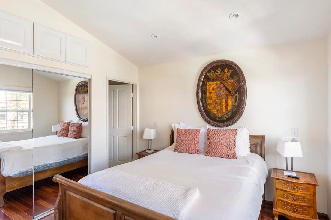 A bedroom with a large bed, a mirror, a wooden nightstand, and a wall-mounted artwork.