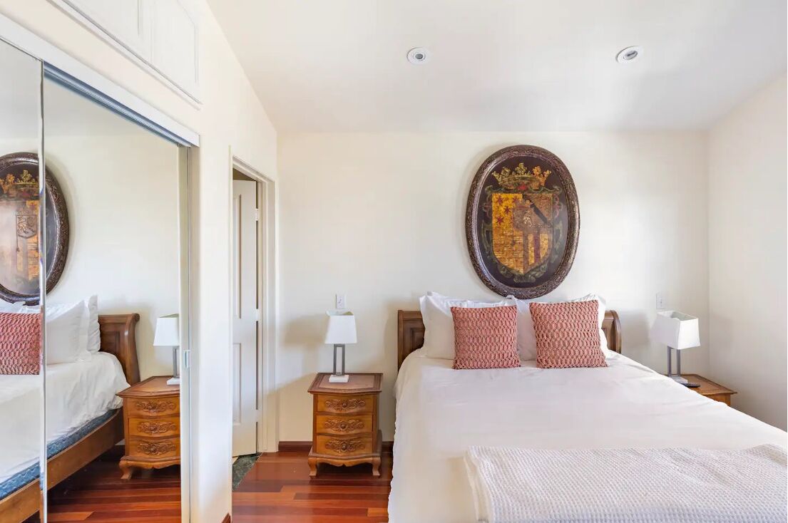 Quarto de hotel com uma cama grande, duas mesas de cabeceira, um espelho e um objeto decorativo na parede.