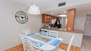 Un comedor con una mesa y sillas blancas, un reloj decorativo y una cocina al fondo.