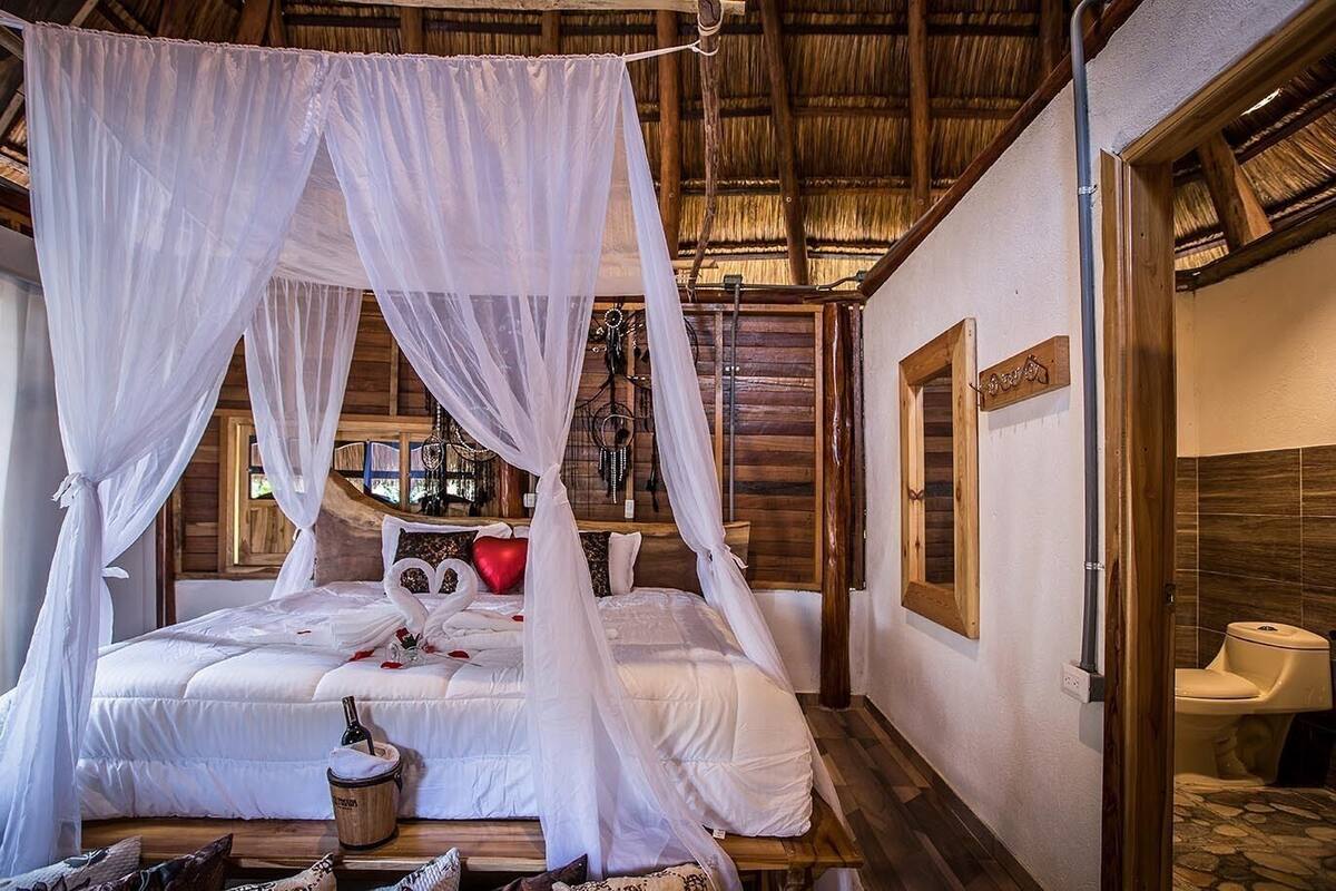A room with a bed under a canopy, a wooden floor, a small table with a bucket, and a toilet.