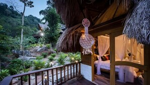 Una terraza de madera con techo de paja, una cama con ropa blanca y vistas a un jardín exuberante.