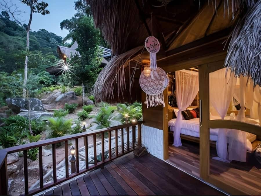 A wooden deck with a thatched roof, a bed with white bedding, and a view of a lush garden.