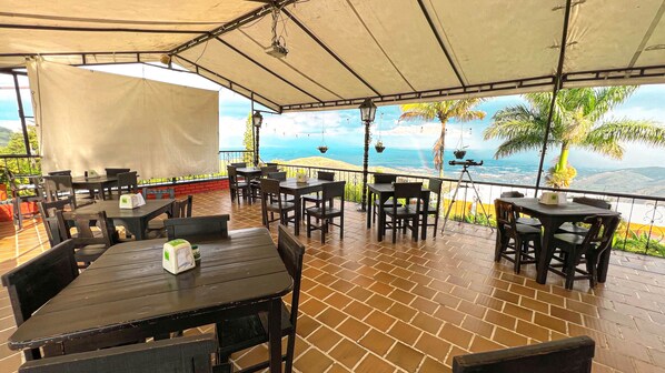 Un patio al aire libre con mesas y sillas, vista al mar y una zona cubierta con toldo blanco.