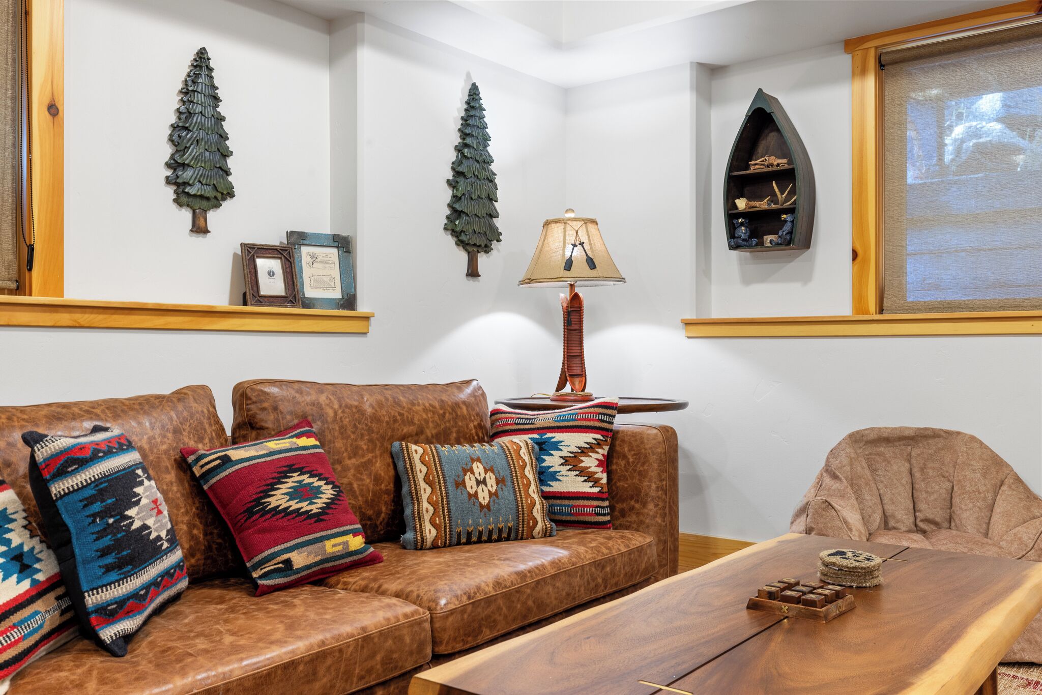 Una sala de estar con un sofá de cuero marrón, almohadones coloridos, una mesa de centro de madera, una lámpara y adornos en la pared, incluyendo dos árboles de Navidad y una estantería con objetos.