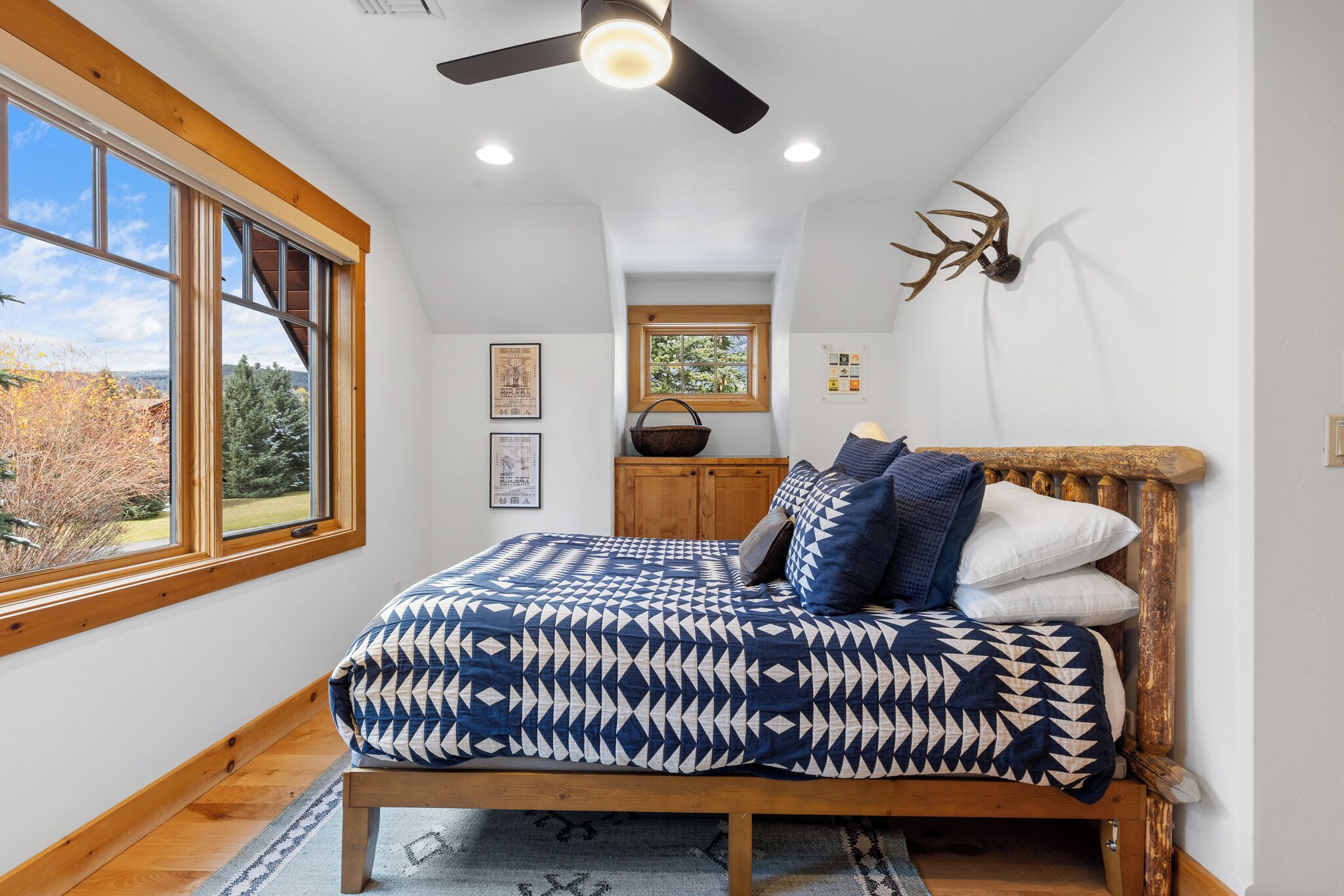 Ein Schlafzimmer mit großem Fenster, Deckenventilator, Bett mit gemusterten Bettbezügen, Holz-Kopfteil und Dekoration aus Hirschgeweihen.