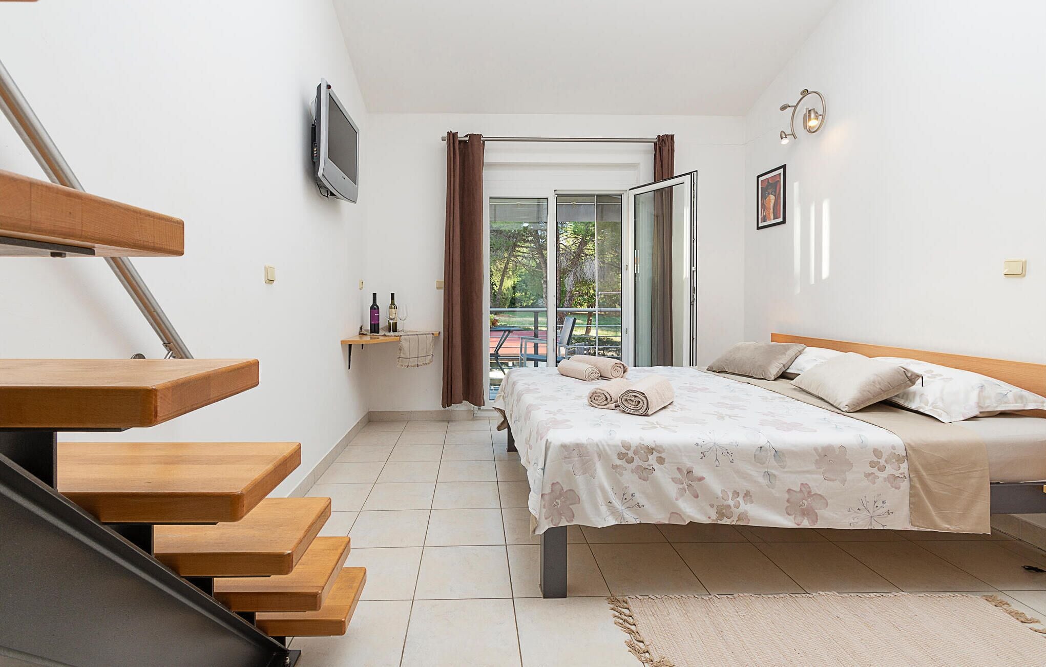 A bedroom with a bed, a desk, a chair, a TV, and a balcony with curtains.