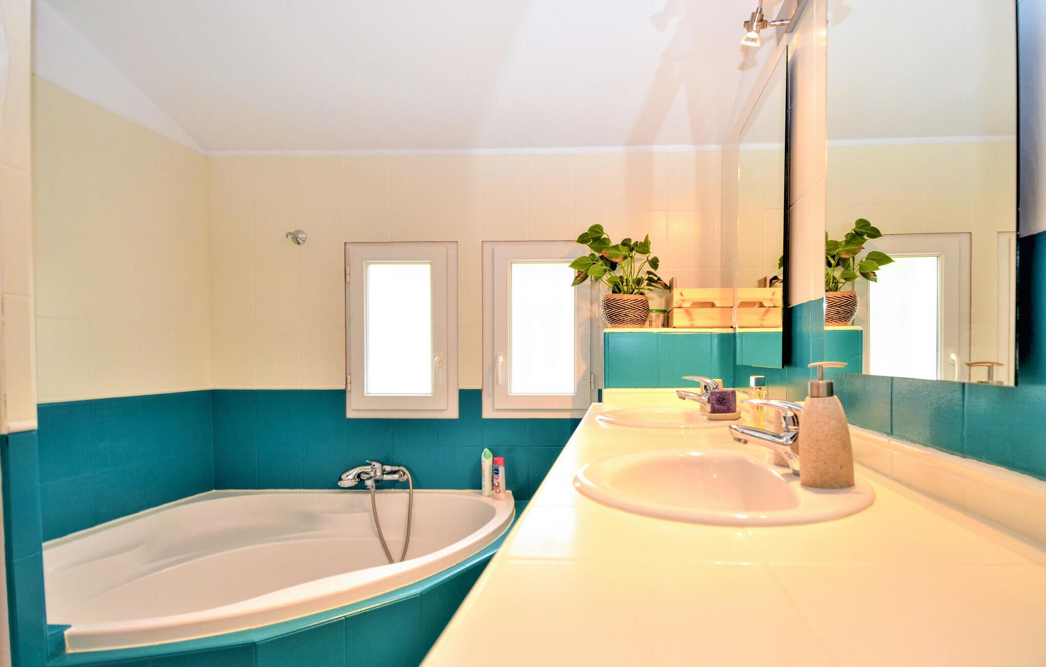 Un baño con bañera, lavamanos, un espejo grande y dos ventanas con plantas.