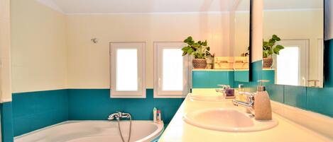 Un baño con bañera, lavamanos, un espejo grande y dos ventanas con plantas.