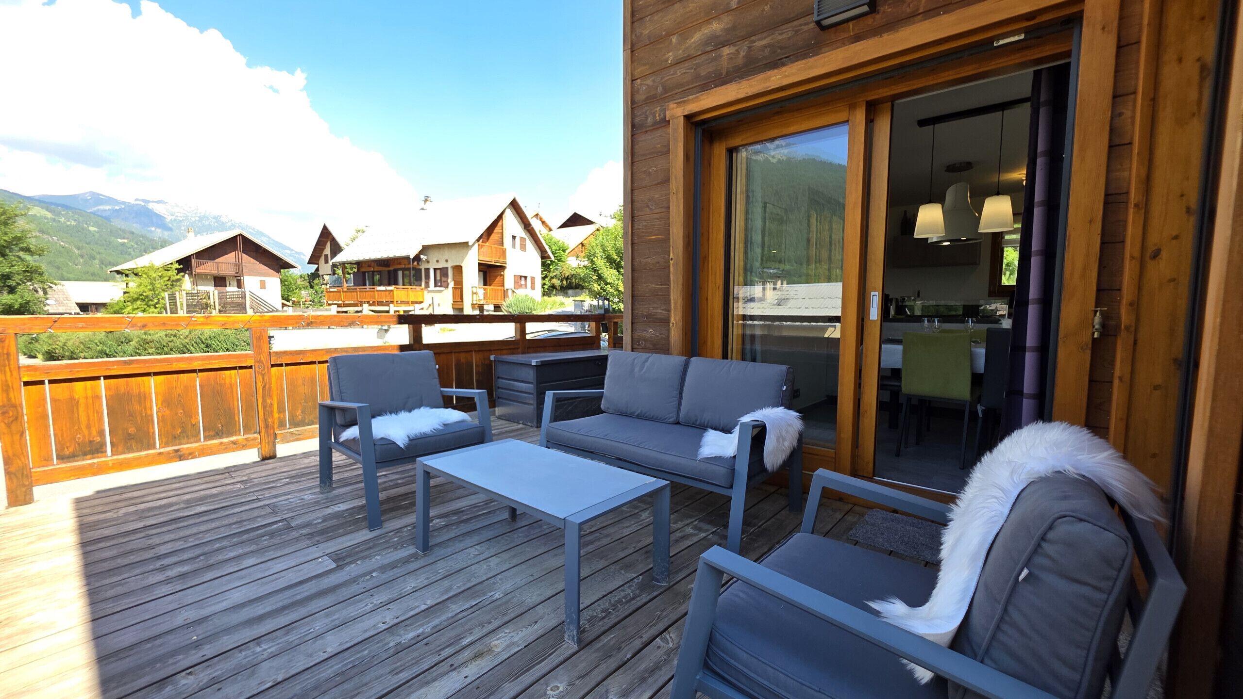 Une terrasse en bois avec des meubles d’extérieur, une porte vitrée donnant sur une salle à manger intérieure et, en arrière-plan, une vue sur les montagnes.