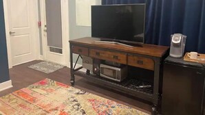 Huoneessa on televisio, joka on sijoitettu puiseen telineeseen, kahvinkeitin ja kuviollinen matto.