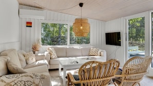 A cozy living room with a white sofa, a coffee table, and wicker chairs. There is a large window with a view of trees, a hanging light fixture, and a flat-screen TV mounted on the wall. - Gorgeous home in Højby with WiFi (Højby)